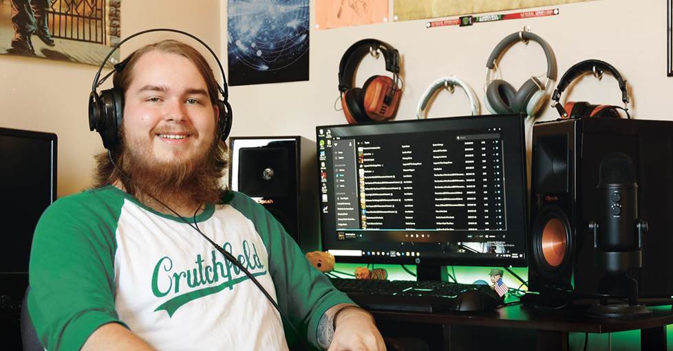 Archer, sitting at his desk at home, with several headphones hanging from hooks on the wall