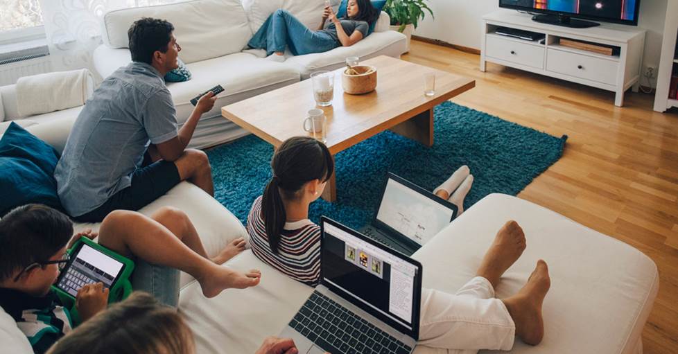 A family in a living room. The adult man is watching tv, the adult female is working on a laptop computer, three kids are using a phones, a tablet, and a laptop