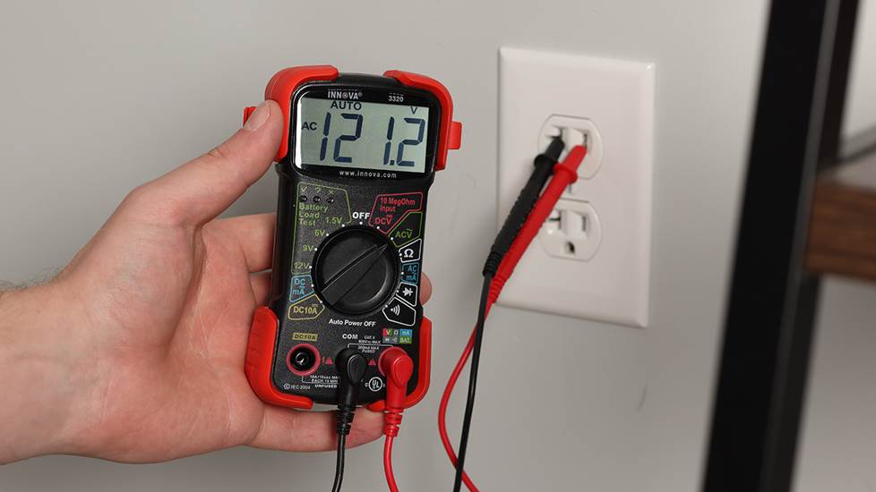 Hand holding a multimeter with probes in wall outlet