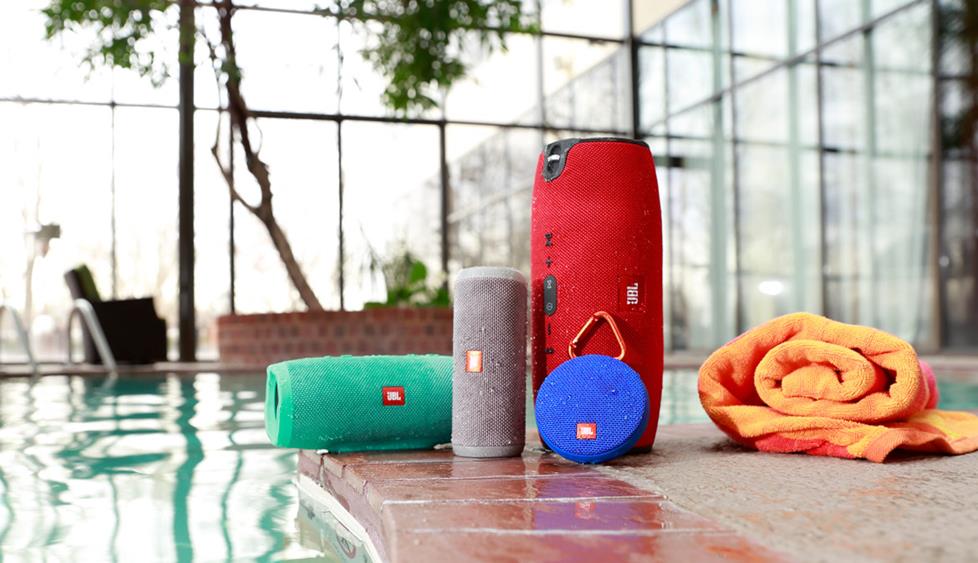 Several bluetooth speakers, in assorted shapes and colors, beside a swimming pool.