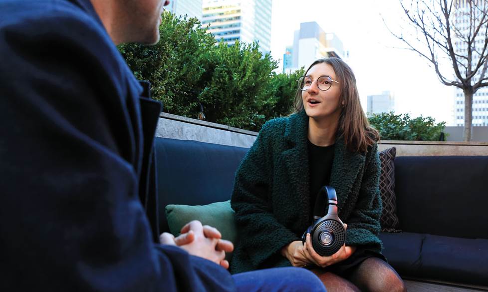 Woman site on a rooftop bench and speaks to a man who's shoulder is seen in the foreground. City buildings appear behind her.