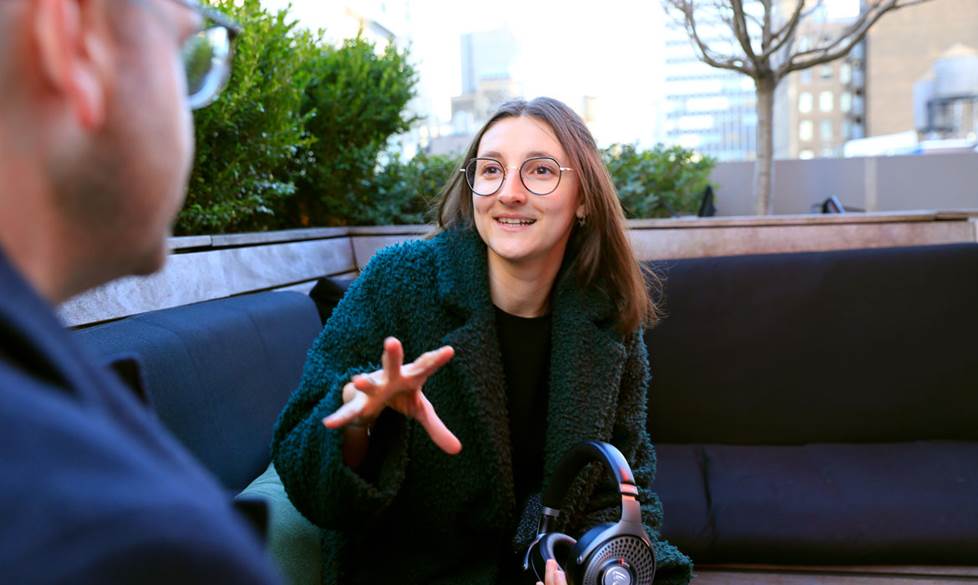 Photo of woman gesturing with her hand. She is sitting on a bench on a rooftop and speaking to a man in the foreground. The city is behind her.