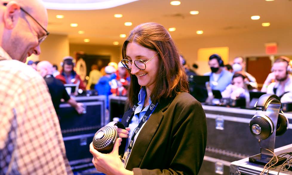 Photo of Megáne standing in the center of the photo with a crowd of people behind her. She is holding a pair of headphones in her hands.