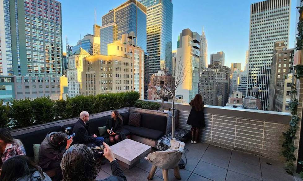 A behind-the-scenes shot of Jeff and Megáne sitting on the rooftop lounge. There are city buildings rising in the background against the blue sky. To the bottom left are photographers who are shooting the scene.