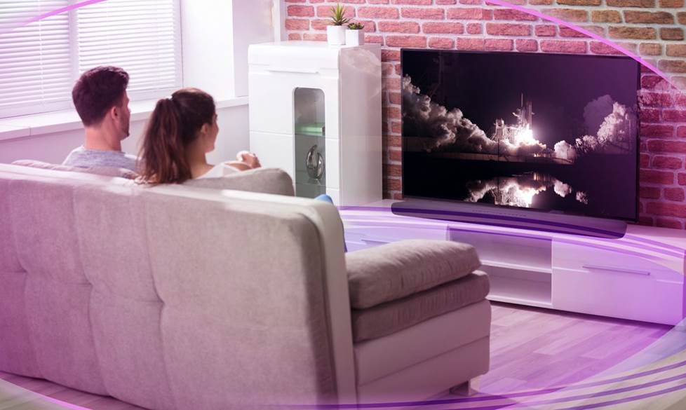 Two people sitting on a couch watching TV while a purple representation of surround sound makes a bubble around them.
